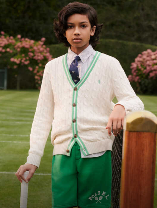 Boy wears a cream cardigan and green shorts while holding a tennis racquet.