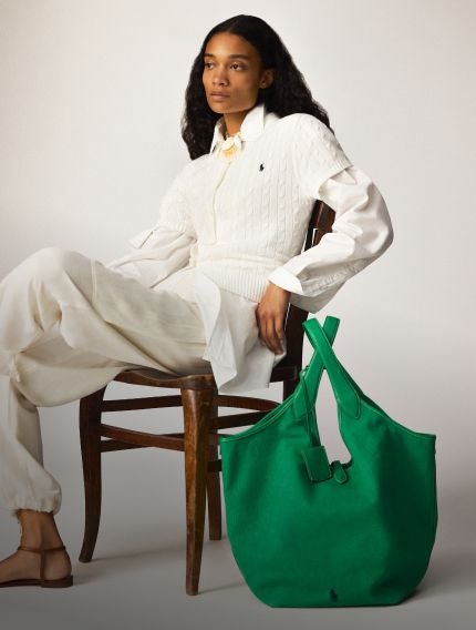 Woman in chair wears all-white outfit next to large green handbag.
