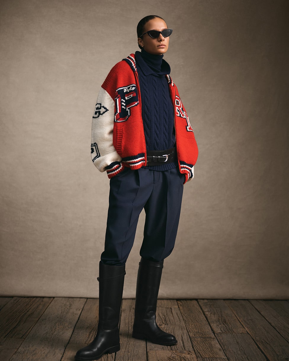 Woman standing wearing a red and white varsity jacket, navy jumper and trousers and tall black boots.